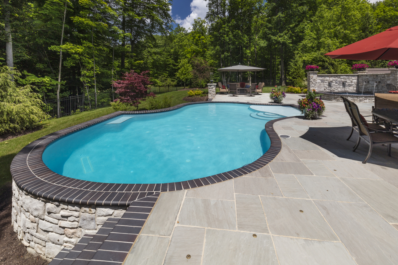 Freeform residential pools installed by a large wooded area.