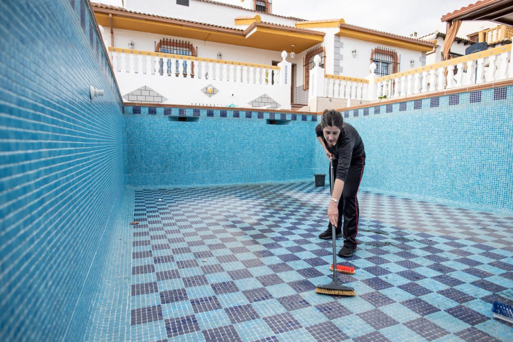 Woman scrubbing the floor of an empty tiled swimming pool.